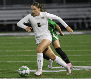 Soccer player in white uniform dribbling ball on field during a competitive match at night.