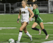Two female soccer players competing in a match under stadium lights; player in white dribbles the ball, closely followed by a defender.