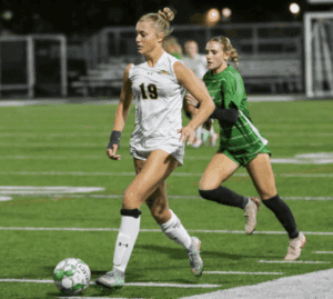 Two female soccer players competing in a match under stadium lights; player in white dribbles the ball, closely followed by a defender.