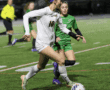 Soccer player in white jersey dribbles ball past opponent on field under lights during a competitive match.