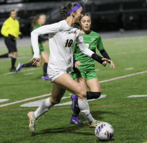 Soccer player in white jersey dribbles ball past opponent on field under lights during a competitive match.