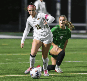 Soccer player in white uniform dribbles a ball on the field, with a green-uniformed opponent closely following.
