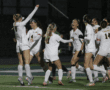 Girls' soccer team celebrating a win under the lights on a rainy field.