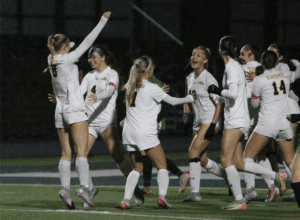 Girls' soccer team celebrating a win under the lights on a rainy field.