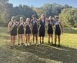 Rowing team posing with medals in front of a scenic park backdrop on a sunny day.