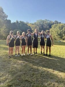 Rowing team posing with medals in front of a scenic park backdrop on a sunny day.