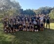Group of smiling rowers wearing medals and team uniforms, posing outdoors on a sunny day.
