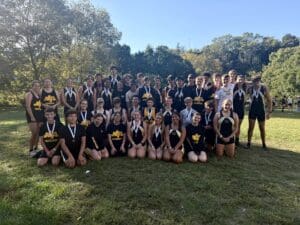 Group of smiling rowers wearing medals and team uniforms, posing outdoors on a sunny day.