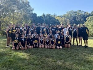 Rowing team group photo in uniforms on sunny day with medals and wooded backdrop.