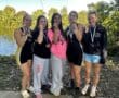 Group of five female athletes by a lake, celebrating with medals, showing team spirit and achievement.