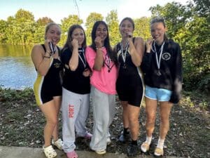 Group of five female athletes by a lake, celebrating with medals, showing team spirit and achievement.
