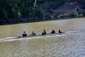 Four-person rowing team in a sleek rowing boat on a river, surrounded by lush greenery in the background.