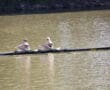 Two rowers in a black boat labeled True Grit on a calm river, competing in a synchronized rowing event.