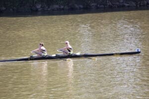 Two rowers in a black boat labeled True Grit on a calm river, competing in a synchronized rowing event.