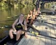 Women's rowing team prepares on sunny river dock, surrounded by trees and bridge.