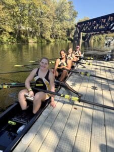 Women's rowing team prepares on sunny river dock, surrounded by trees and bridge.