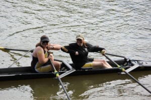 Two women rowing a boat on a river, smiling, and wearing matching athletic outfits and caps.