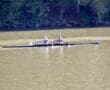 Two people rowing in a double scull boat on a calm river, surrounded by trees.