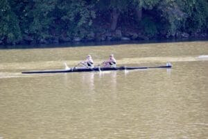 Two people rowing in a double scull boat on a calm river, surrounded by trees.