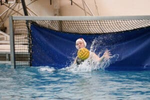 Water polo goalkeeper blocking ball in pool goal with splashing water.