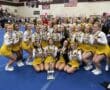 Cheerleading team in yellow uniforms celebrating with a trophy in a gymnasium.