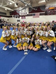 Cheerleading team in yellow uniforms celebrating with a trophy in a gymnasium.