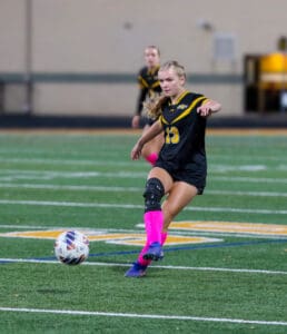 Soccer player in action kicking ball, wearing a black uniform and pink socks on a green field.