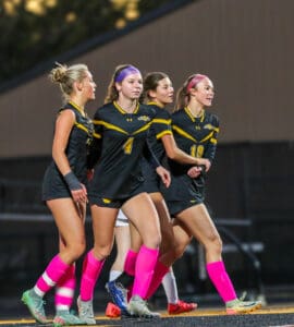 Four female soccer players in black and yellow jerseys celebrate on the field, wearing bright pink socks.