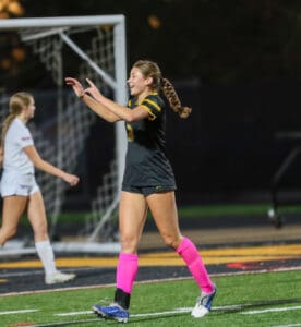 Soccer player in black uniform celebrates on the field, wearing pink socks, with a teammate nearby during a night game.