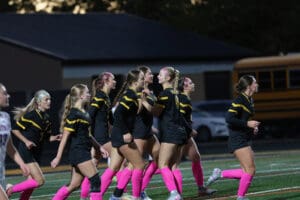 Girls' soccer team celebrates on field in black uniforms and pink socks during a night match.