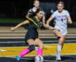Soccer player in black controls the ball, challenged by opponents in white, during an intense night match on the field.