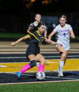 Soccer player in black controls the ball, challenged by opponents in white, during an intense night match on the field.