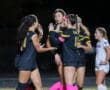 Soccer team celebrates with high-fives during a night game, wearing black jerseys and pink accessories.