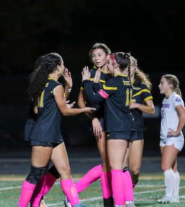 Soccer team celebrates with high-fives during a night game, wearing black jerseys and pink accessories.