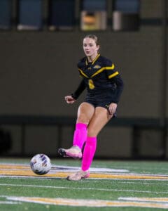 Soccer player in black and yellow uniform, pink socks, kicking ball on field at night.