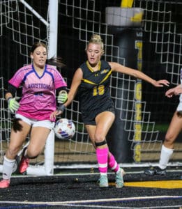 Soccer player in black uniform and goalie competing for ball near goal net during intense match.