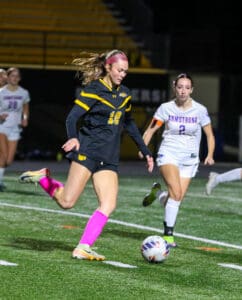 Soccer match action with a player in black and yellow kicking the ball, pursued by an opposing player in white.