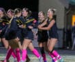 Women's soccer team celebrates on field, wearing black uniforms with pink socks during a night game.