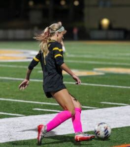 Female soccer player in black and yellow uniform kicking ball on field at night.