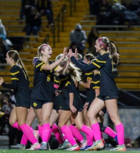 Girls' soccer team celebrates with high-fives, wearing black uniforms with pink socks on the field at night.