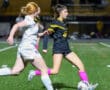 Two female soccer players from opposing teams compete for the ball during a match on a field at night.