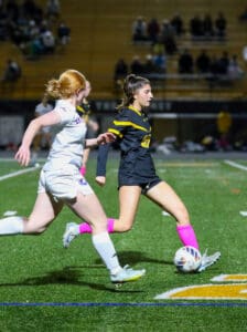 Two female soccer players from opposing teams compete for the ball during a match on a field at night.