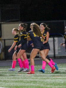 Women soccer players celebrate on the field wearing pink socks and dark uniforms at night.