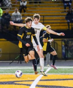 Soccer match action with two players competing for the ball on a field with orange and black markings.