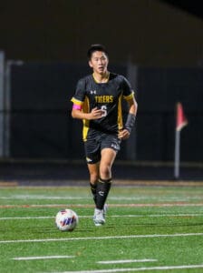 Soccer player in black uniform dribbling ball on field at night, action shot with corner flag in background.