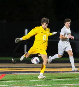 Soccer goalie in yellow uniform kicks the ball during a nighttime match on a green field, teammate in white nearby.