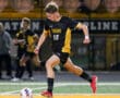 Soccer player in black and gold uniform kicks ball on field during match.