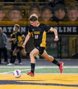 Soccer player in black and gold uniform kicks ball on field during match.