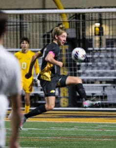 Soccer player in black uniform, mid-action on the field, focused with a soccer ball in motion nearby.