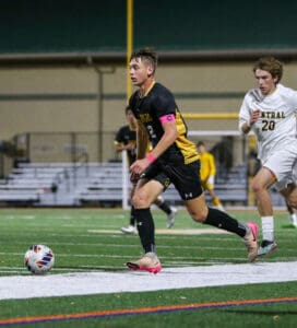 Soccer player in black and yellow uniform dribbles ball during a night match, followed by opponent in white.